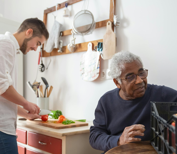 to people in kitchen with one looking into a box and other cutting veg